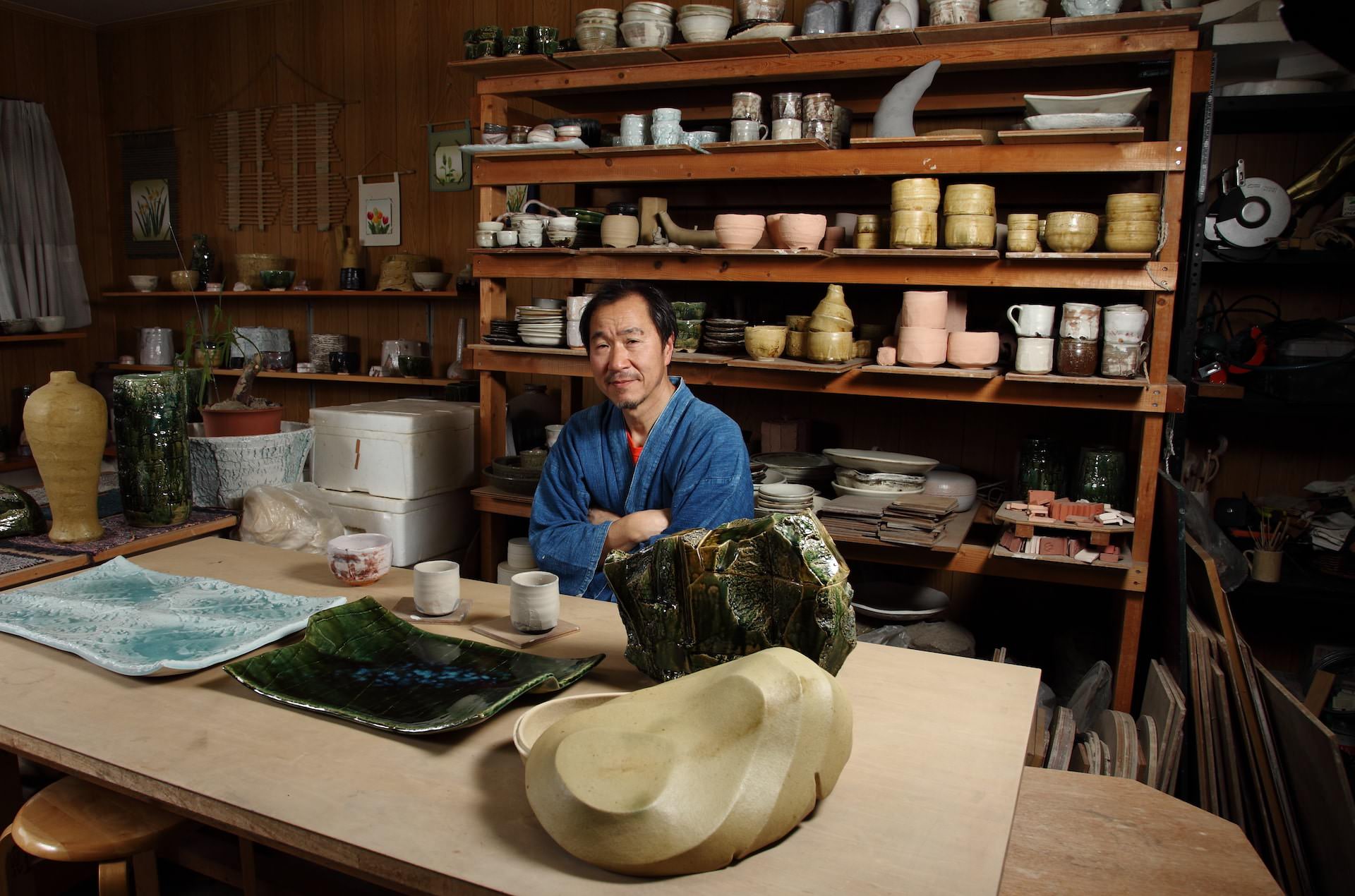 Ceramic artist seated in a workshop surrounded by shelves of handcrafted pottery and ceramic vessels