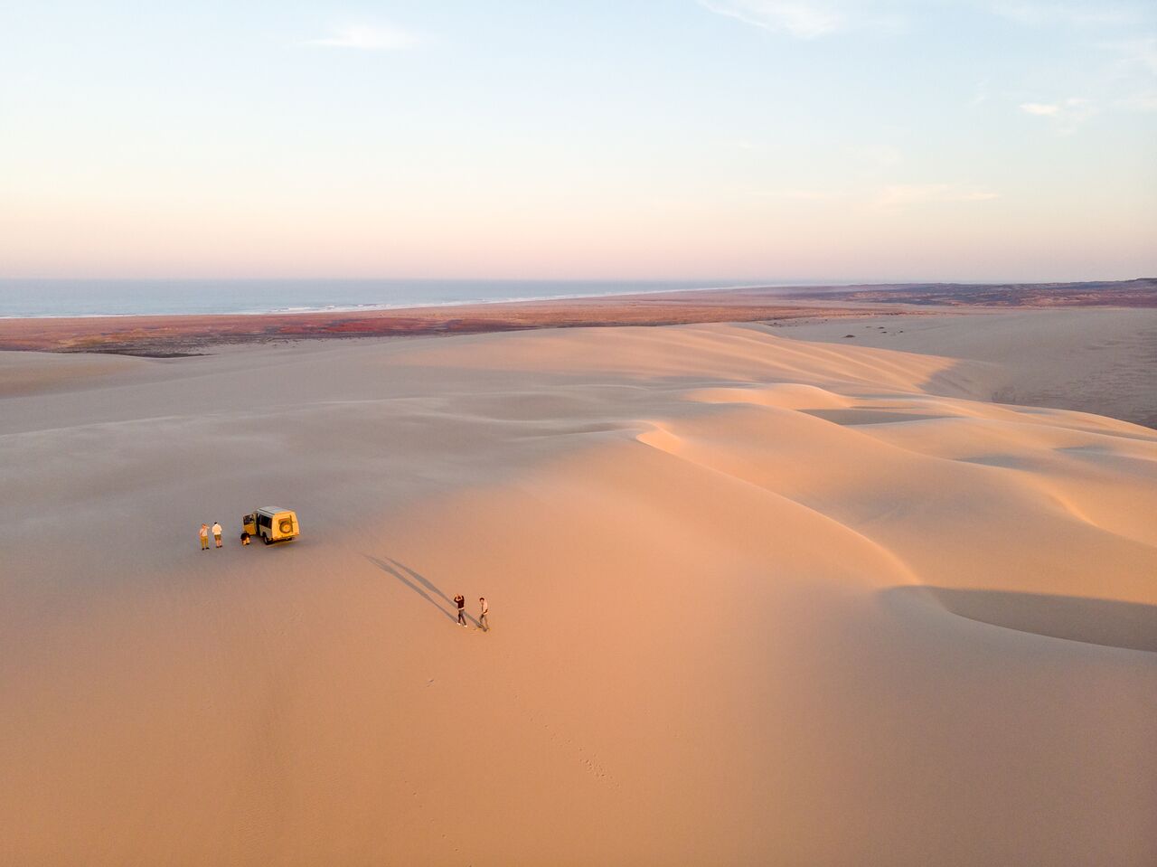 Aerial view of sweeping golden sand dunes along Namibia’s remote coastline, with a safari vehicle and travelers exploring the vast desert landscape beneath soft pastel light at sunrise or sunset.