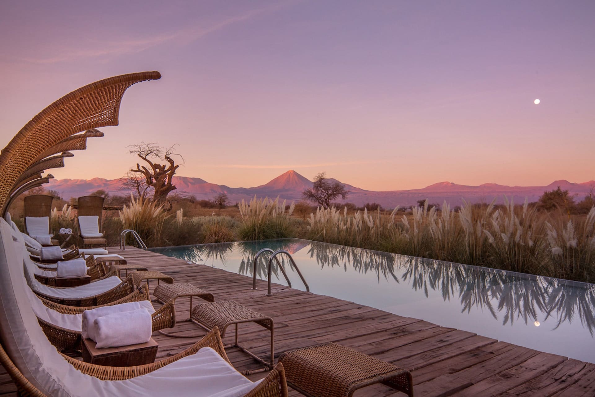 Luxury infinity pool at dusk in Chile’s Atacama Desert, framed by woven lounge chairs and pampas grass, with dramatic volcanic peaks and a glowing pastel sky reflected across still water.