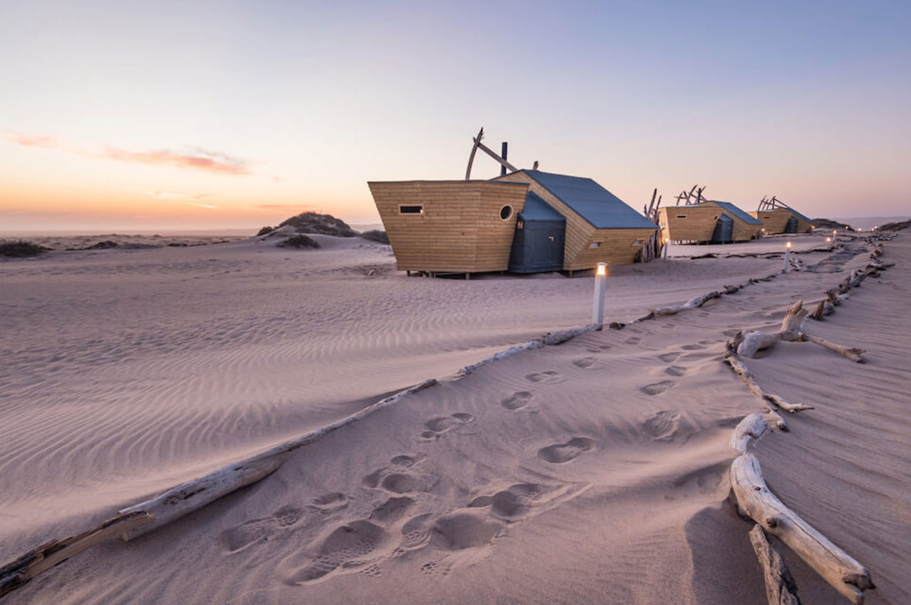 Luxury desert cabins at sunset in Namibia, set among rolling sand dunes with weathered driftwood-lined pathways and soft footprints tracing through the sand beneath a pastel evening sky.