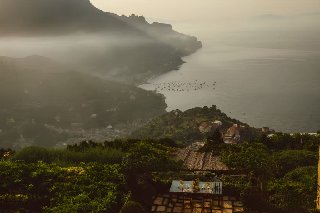 Aerial view of the Amalfi Coast at golden hour with misty mountains, terraced hillside villages, boats in a calm bay, and a candlelit outdoor dining terrace in the foreground