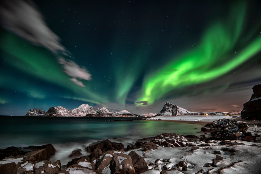 Green northern lights sweeping across a star-filled sky above snowy mountains and a coastal fjord landscape