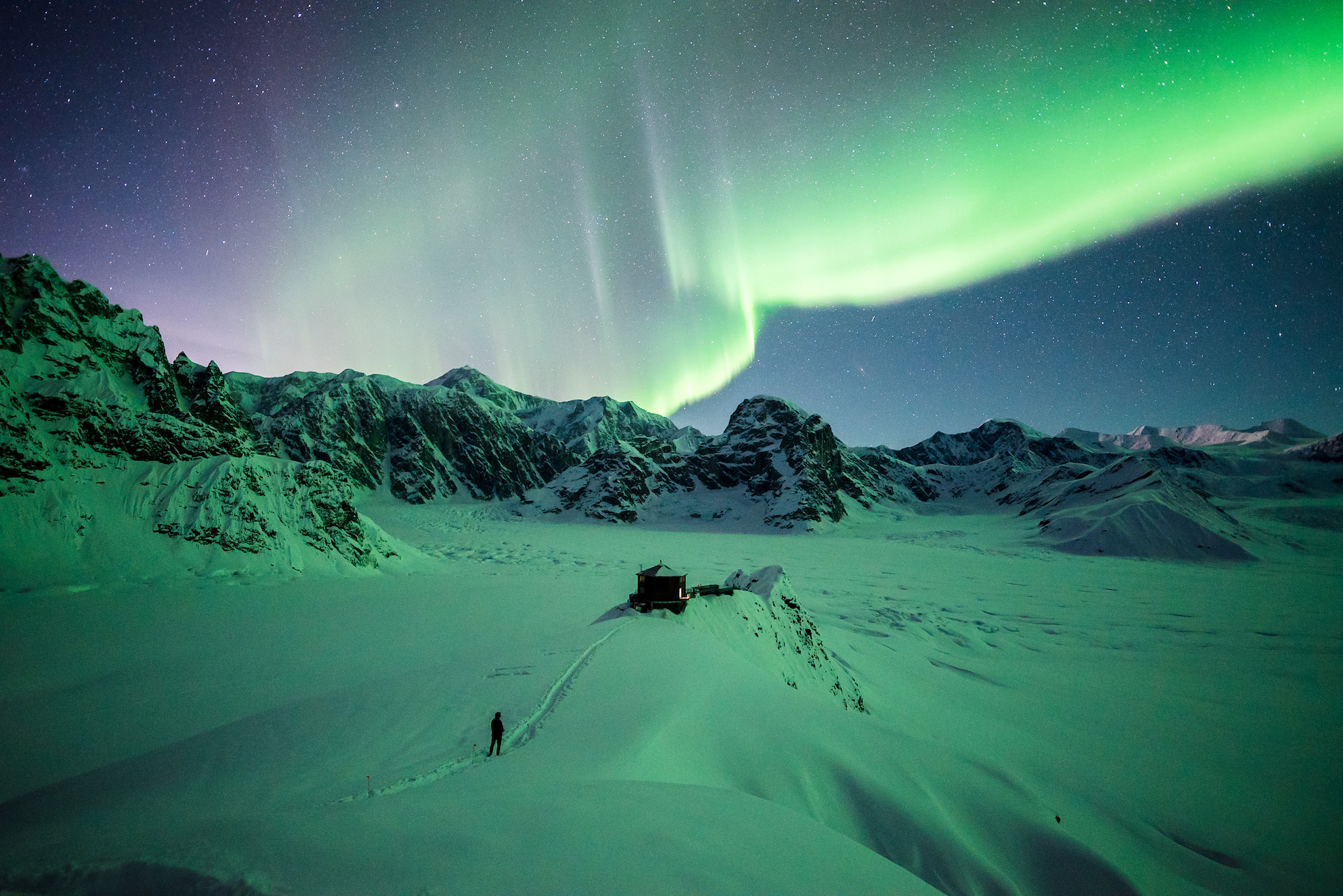 Northern lights above a remote mountain chalet in snow-covered Alaska wilderness with dramatic peaks under a star-filled sky