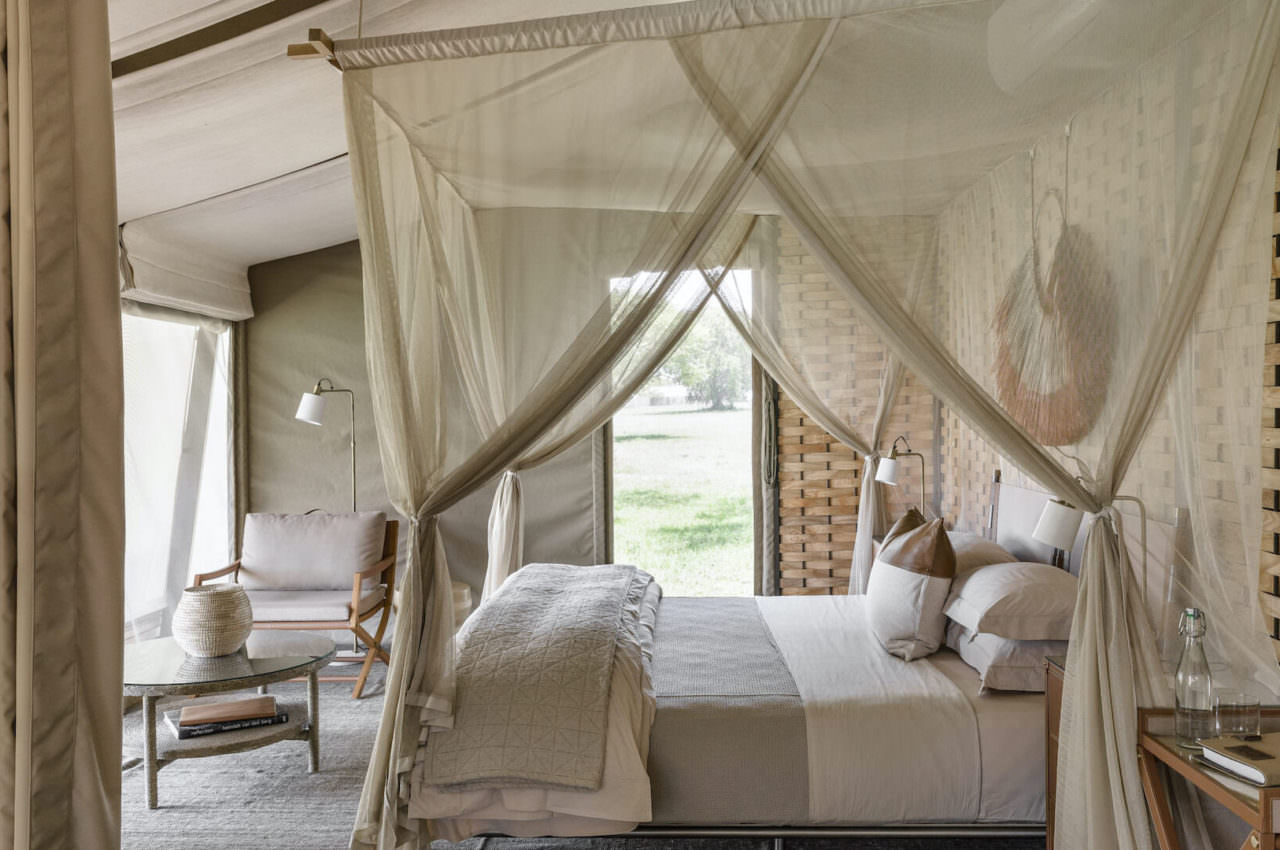 Tented Suite bedroom with sheer canopy four-poster bed, neutral linen bedding, woven wall panels, and open views across the Serengeti plains