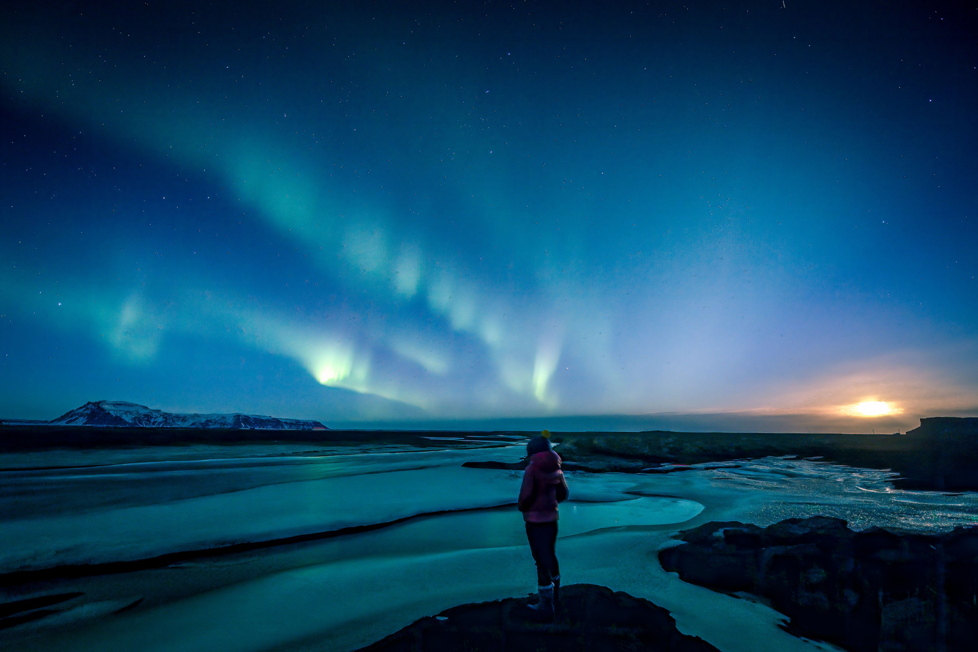Northern lights shimmering above an icy lagoon with a traveler standing on dark rocks under a star-filled sky