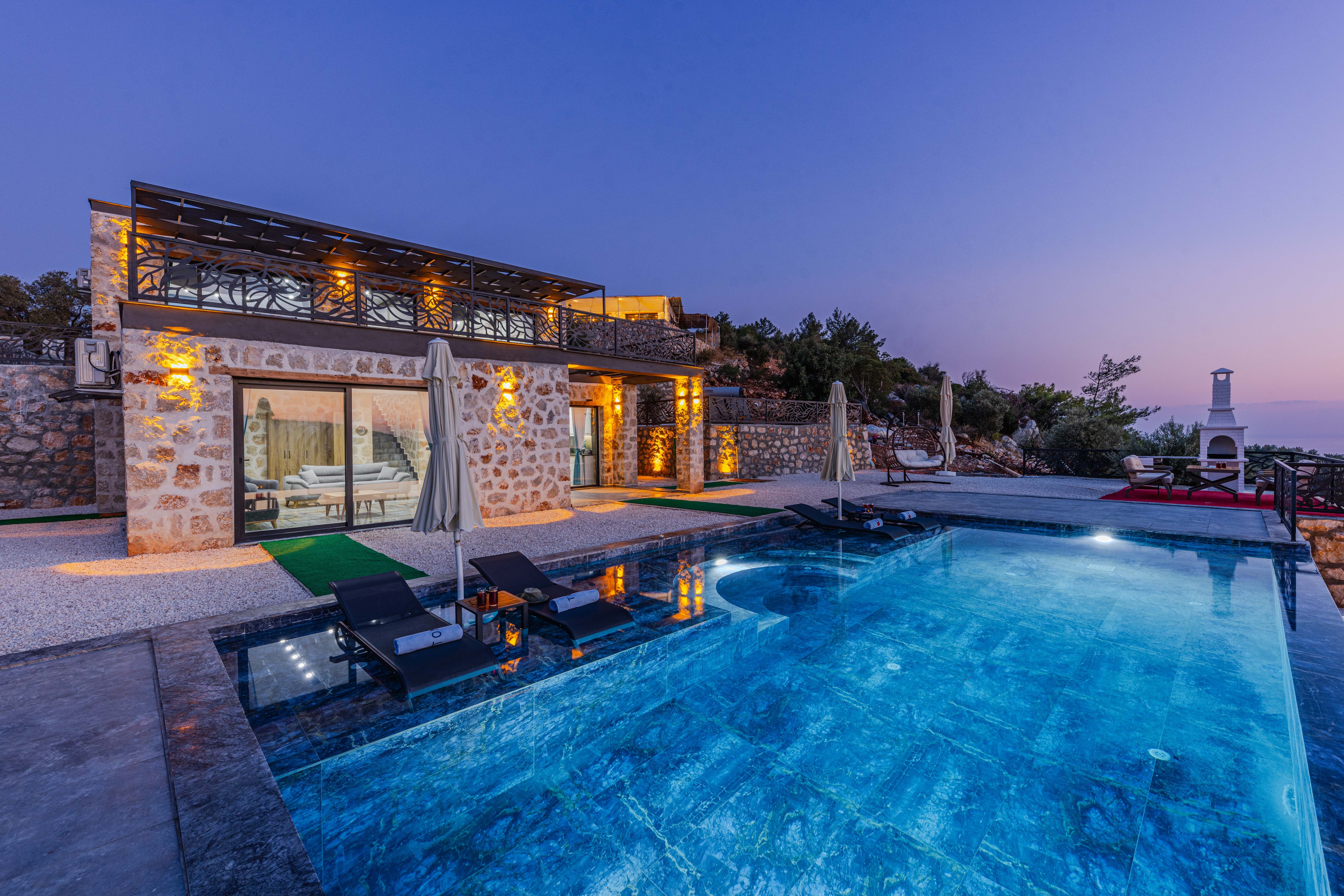 Luxury stone villa at dusk with illuminated infinity pool, sun loungers, and ornate balcony overlooking a hillside Mediterranean landscape