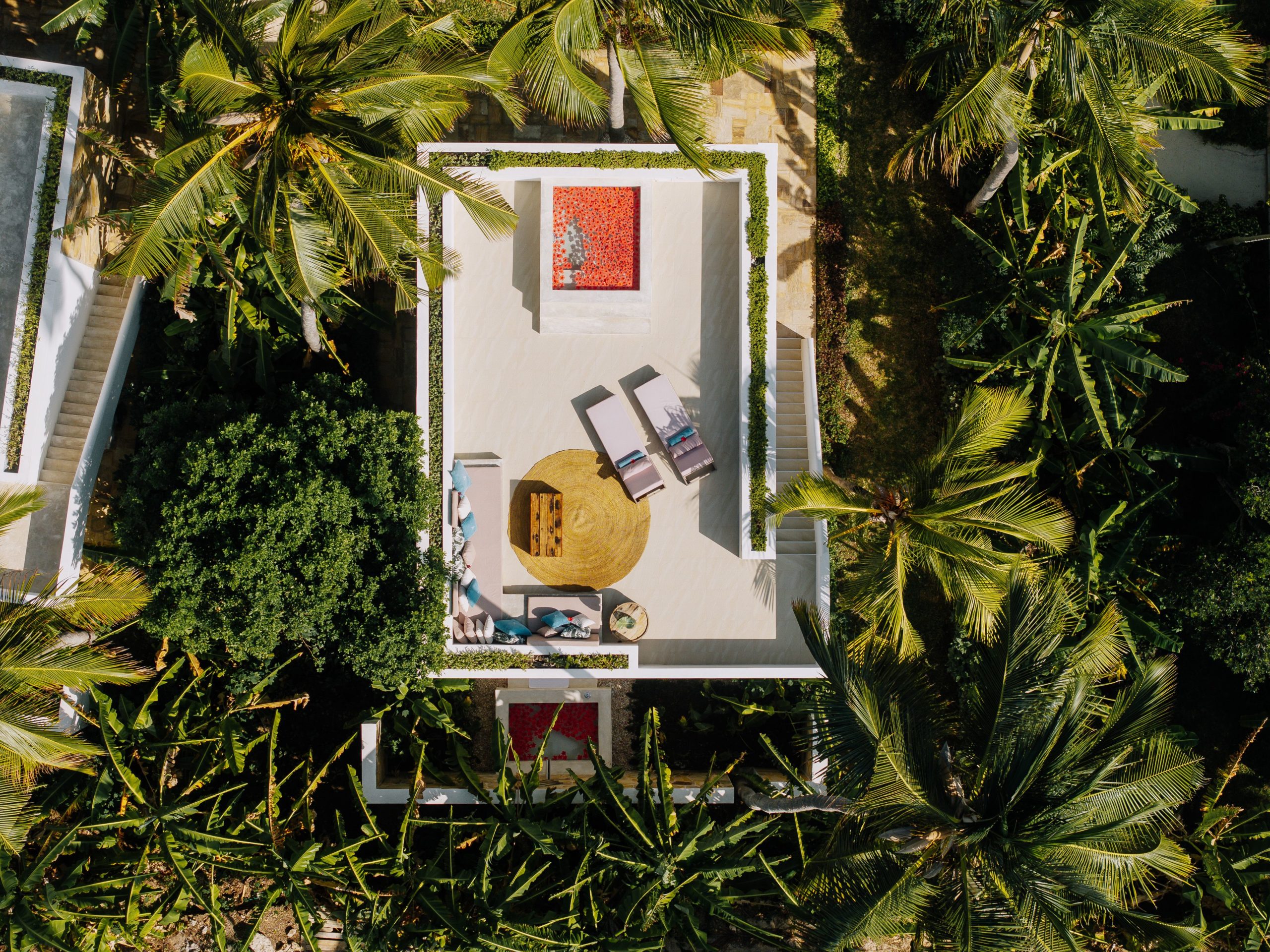 Aerial view of a luxury private villa terrace with sun loungers, circular rattan rug, and vibrant artwork, surrounded by tropical palm and banana trees