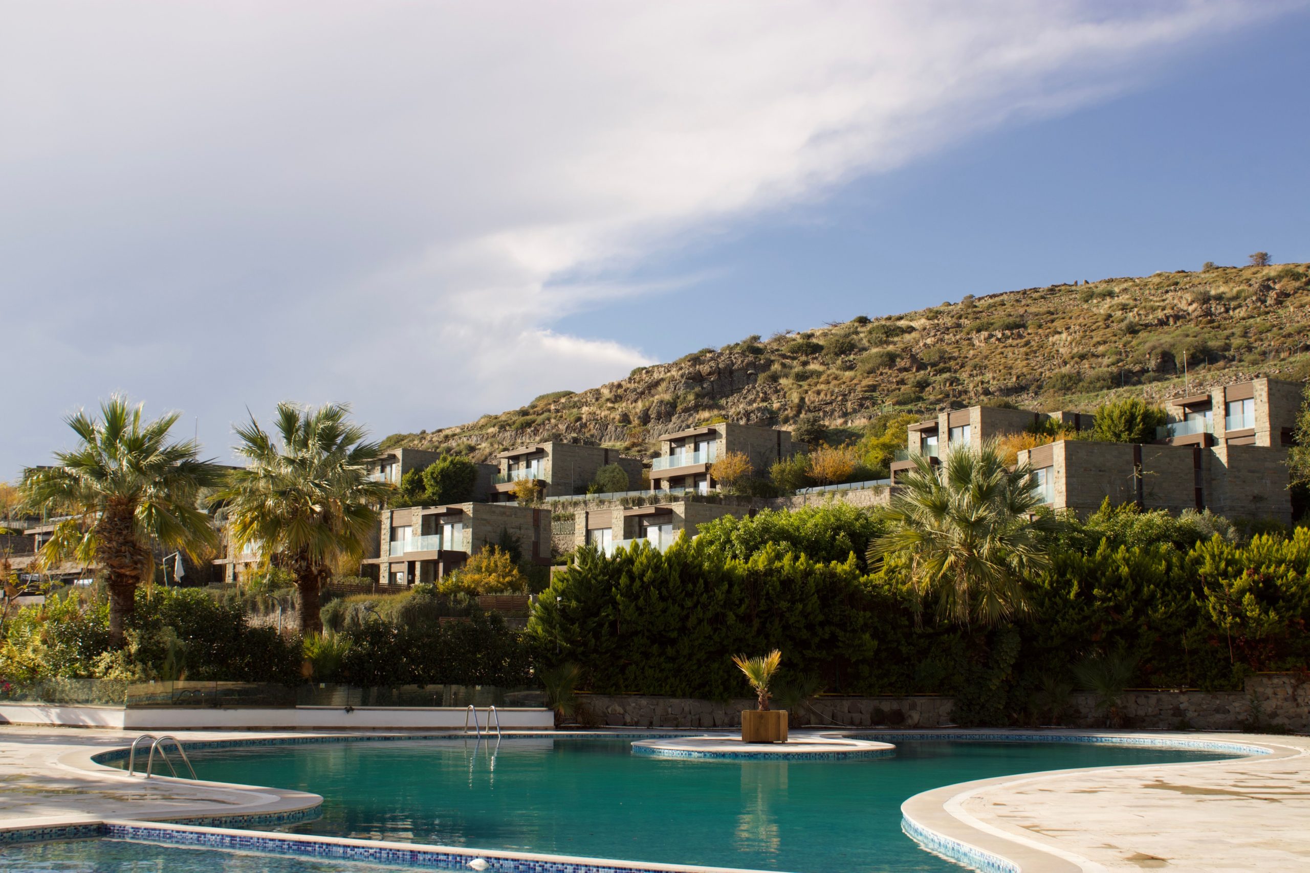Luxury hillside villa resort with tiered stone architecture, glass balconies, and a curved outdoor pool surrounded by palm trees and Mediterranean landscaping