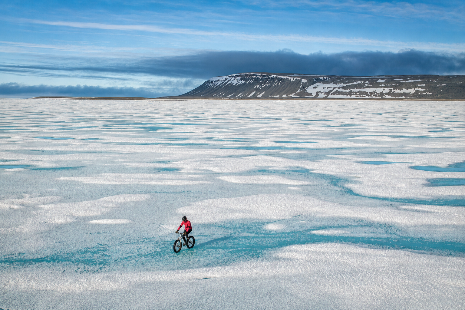 Cyclist riding a fat-tire bike across Arctic sea ice streaked with pale turquoise meltwater, with a snow-dusted mountain range in the distance.
