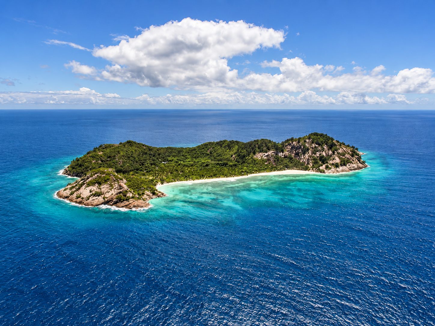 Aerial view of a granite island ringed by white sand and turquoise reef in the Seychelles